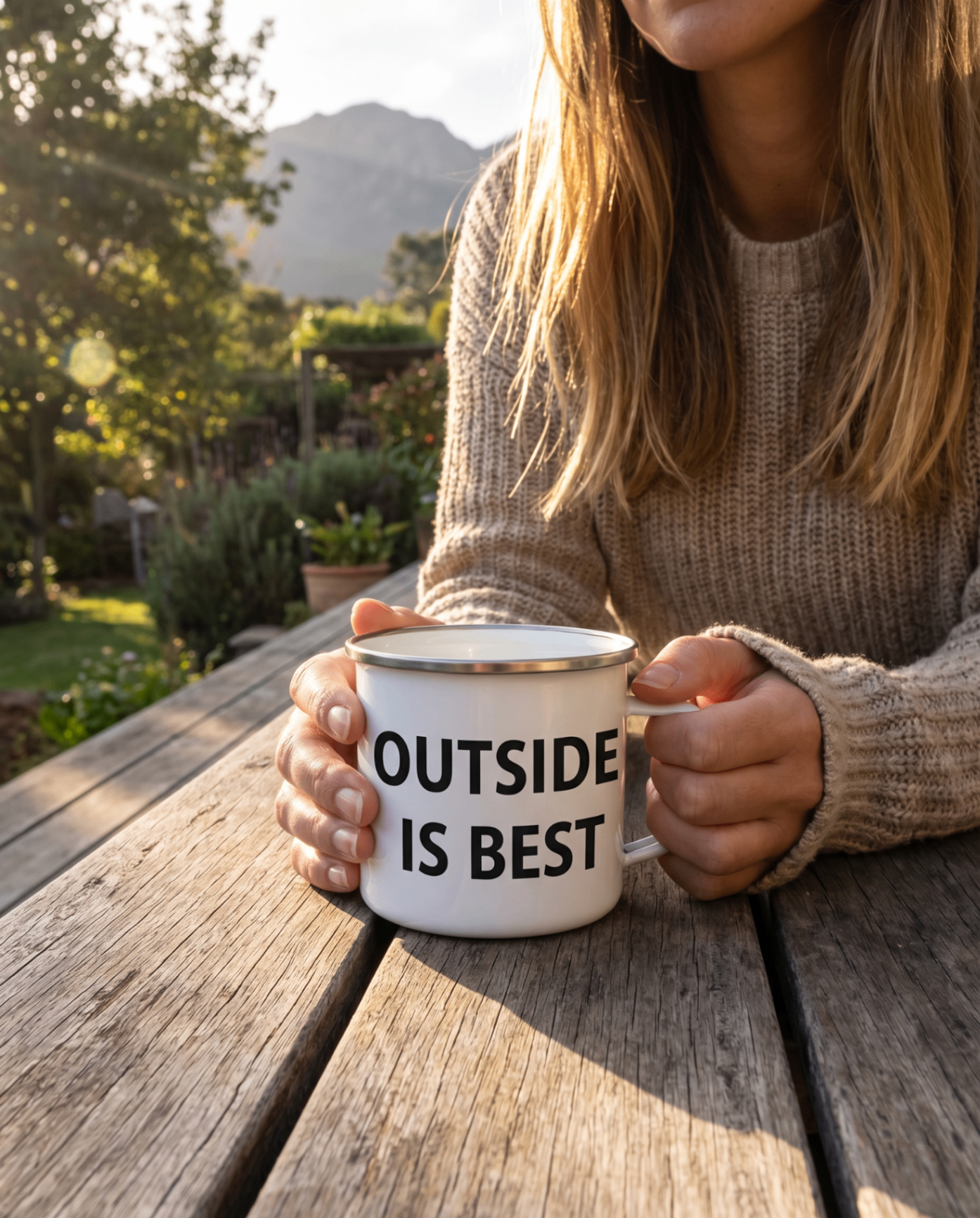 Person in cream sweater holding the Outside Is Best enamel mug at a wooden garden table at golden hour